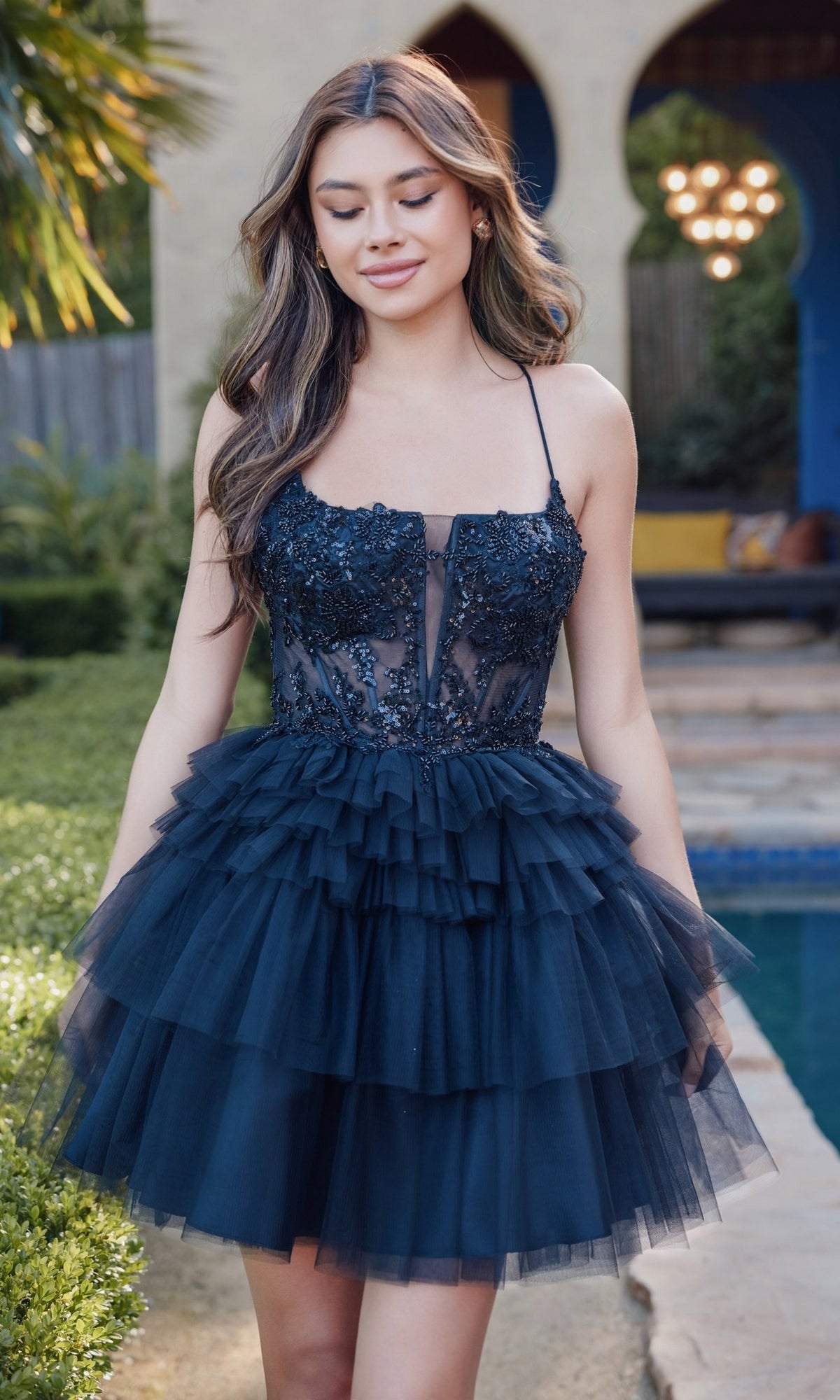 A woman stands outdoors by a pool, wearing the Juliet JT935K Short Ruffled Homecoming Dress with a sleeveless black lace bodice, stylish lace-up back, and layered tulle skirt.